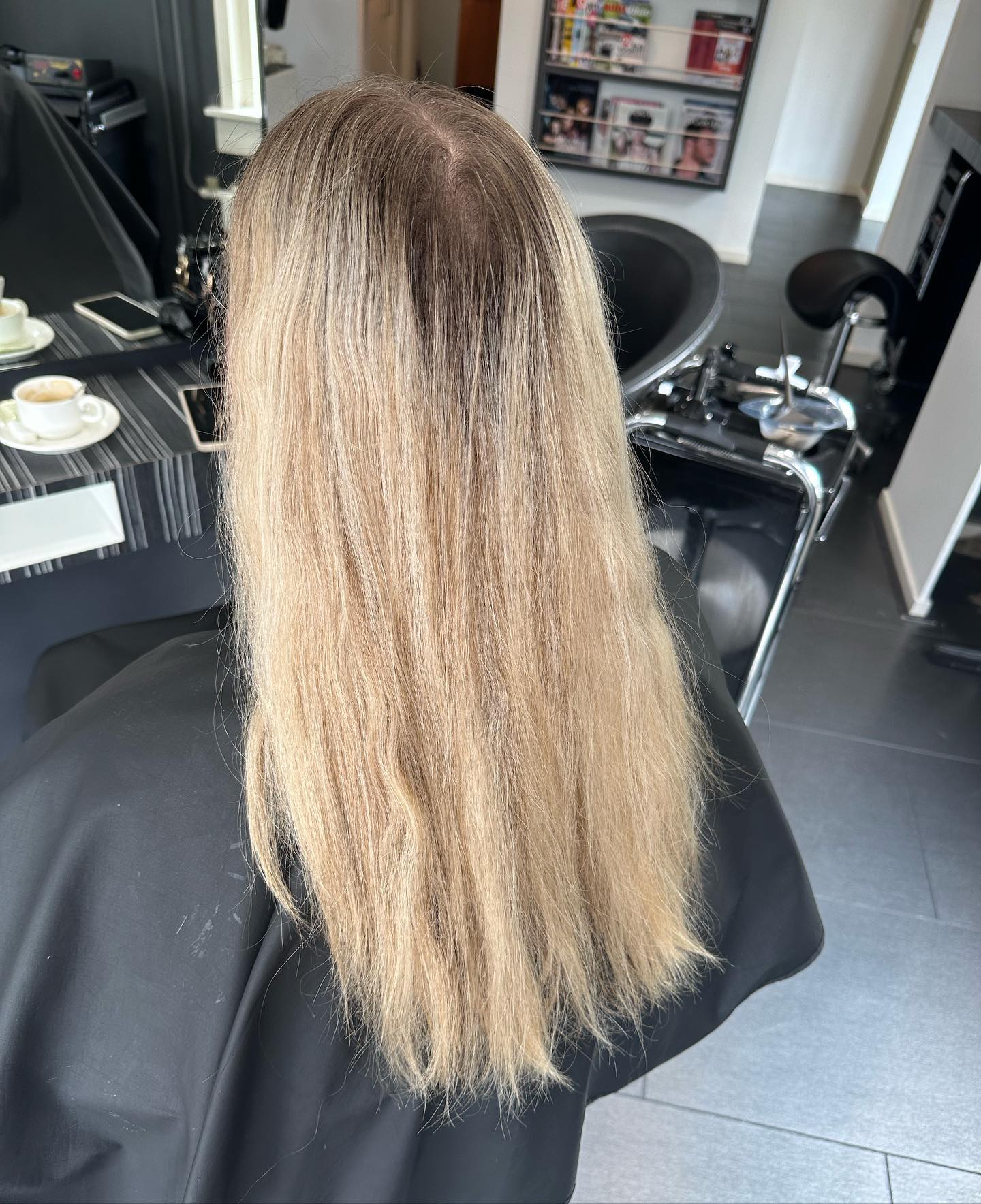 Lang blond haar van achteren in de salon, met een kop koffie op het werkblad en de wasbak op de achtergrond